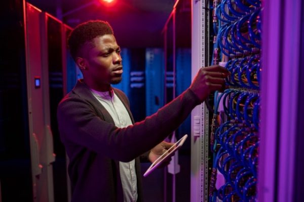 Concentrated young Black man using tablet while examining datacenter equipment in dark server room